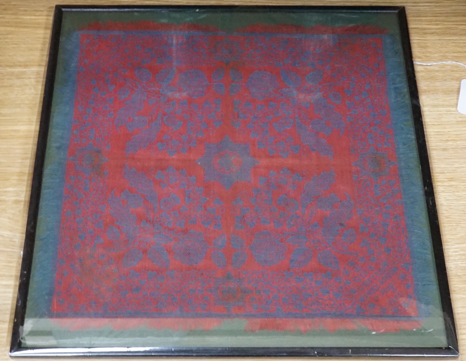 An unusual framed George IV Irish damask napkin, designed with a red ground and blue woven floral design, incorporating Shamrocks, Thistles and Roses, with a cartouche in each corner commemorating King George IV’s visit